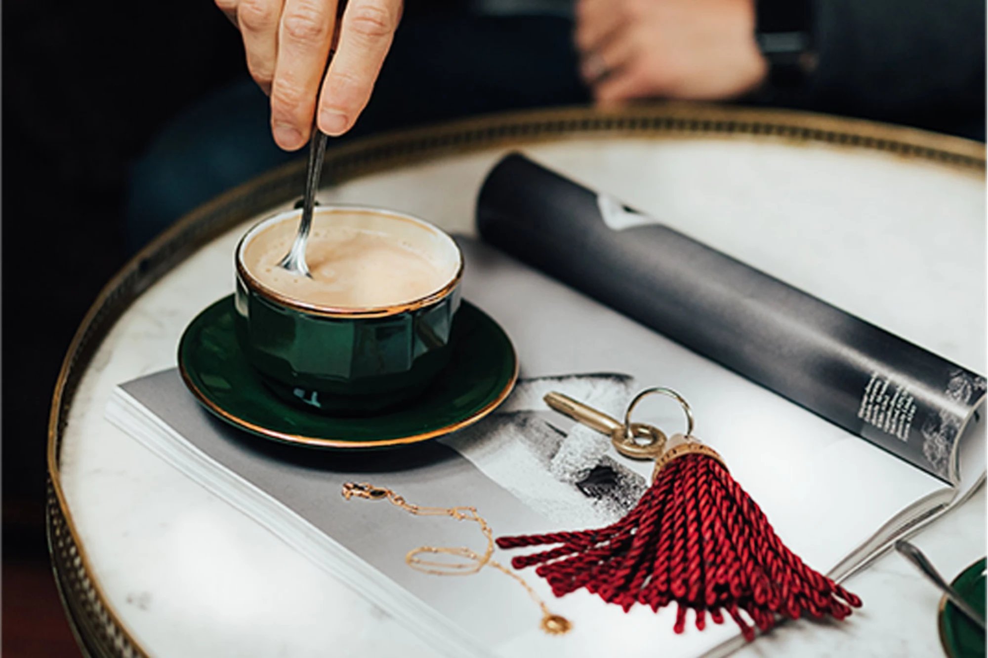 Main remuant une tasse de café vert et or posée sur un guéridon en marbre, à côté d'un magazine